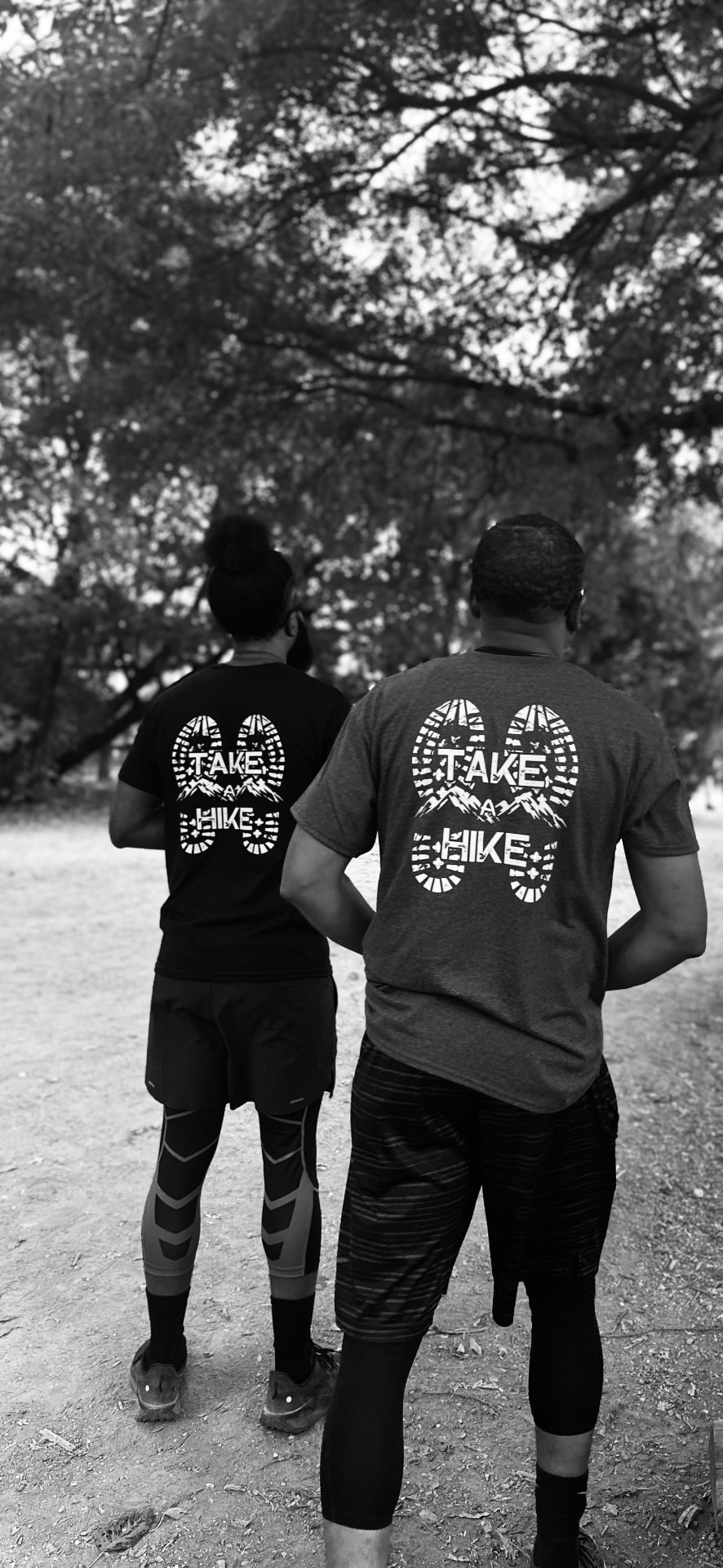 Two hikers from behind wearing black-and-white TAKE A HIKE apparel on a wooded trail